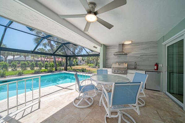 An outdoor patio with a pool, a table with chairs, a ceiling fan, and a kitchen area with a grill.