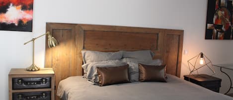 A bedroom with a wooden headboard, a bed with patterned bedding, a bedside table with a lamp, and a wall-mounted picture.