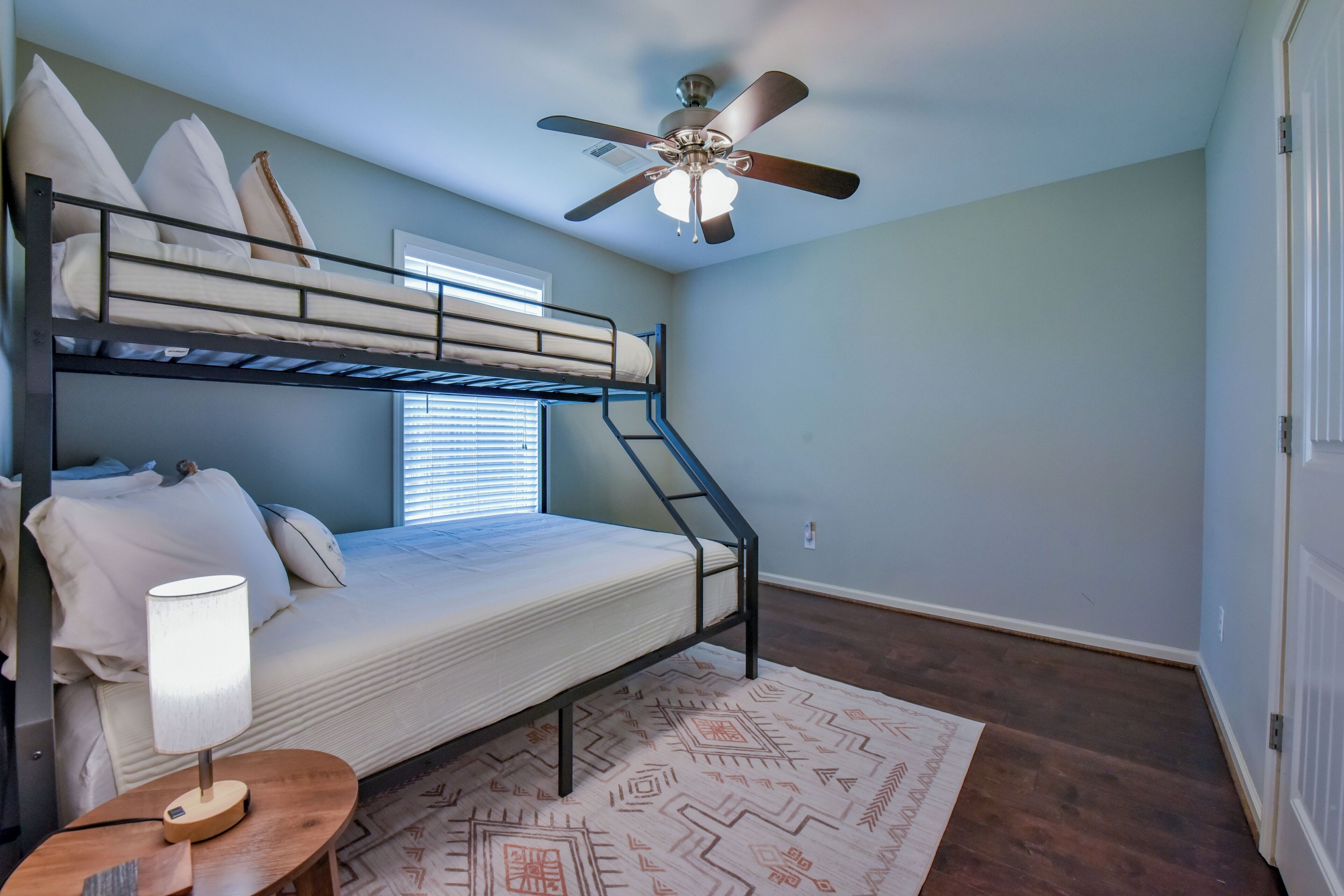 A bunk bed room with a ceiling fan, a window with blinds, a patterned rug, and a bedside table with a lamp.