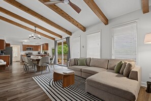 Un salon moderne avec un canapé d’angle, un coin repas avec une table et des chaises, et une cuisine en arrière-plan.