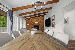 Une salle à manger avec une grande table en bois, des chaises blanches, un lustre, une cheminée en briques et un téléviseur fixé au mur.