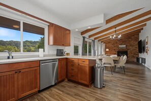 A modern kitchen with wooden cabinets, a stainless steel dishwasher, and a dining area with a brick wall and chandelier. - Galena Home with Golf Course View (Galena)