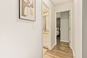 Un couloir avec un mur blanc, une œuvre d’art abstraite encadrée, une salle de bain avec un miroir et un plancher en bois.