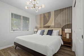 Une chambre moderne avec un grand lit, un luminaire au plafond, une fenêtre avec des stores et un mur orné d’un motif géométrique.