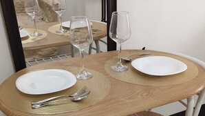 A round dining table with two white plates, two wine glasses, and a set of cutlery. The table has a woven placemat and is supported by a metal frame.