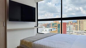 A hotel room with a bed, a TV mounted on the wall, and a large window offering a city view. - NOK Spectacular Loft in Usaqu n (Bogotá)