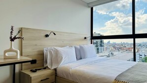 A modern bedroom with a wooden headboard, white bedding, a bedside table with a vase of flowers, and a city view through large windows.