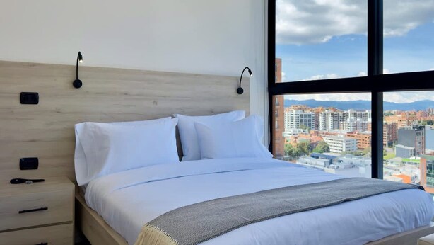 Une chambre avec une grande fenêtre, une tête de lit en bois, un lit recouvert de draps blancs et une vue sur le paysage urbain.