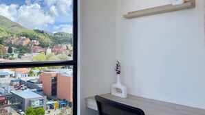 Une pièce dotée d’une grande fenêtre donnant sur un paysage urbain et des montagnes. On y trouve un bureau avec une chaise, ainsi qu’un vase blanc orné de fleurs.