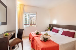 Ein Hotelzimmer mit Schreibtisch, zwei Betten, einem Sessel, einem Fenster mit Vorhängen und Blick auf Gebäude.
