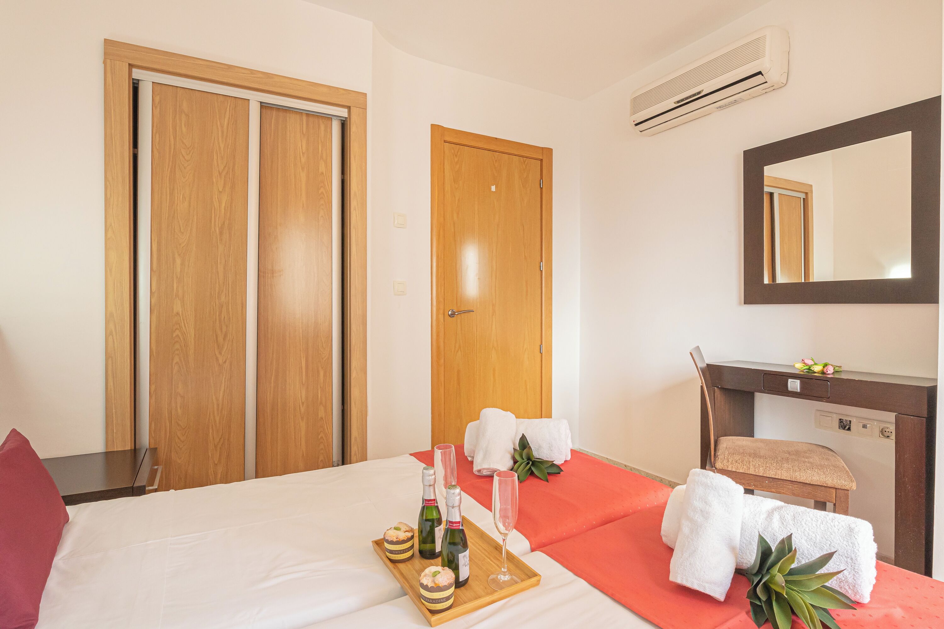 A hotel room with a bed, a wooden bedside table, a tray with bottles and glasses, a chair, and a mirror.
