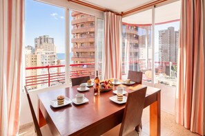 Un coin repas avec une table dressée pour deux personnes, une bouteille d’eau et un panier de fruits.