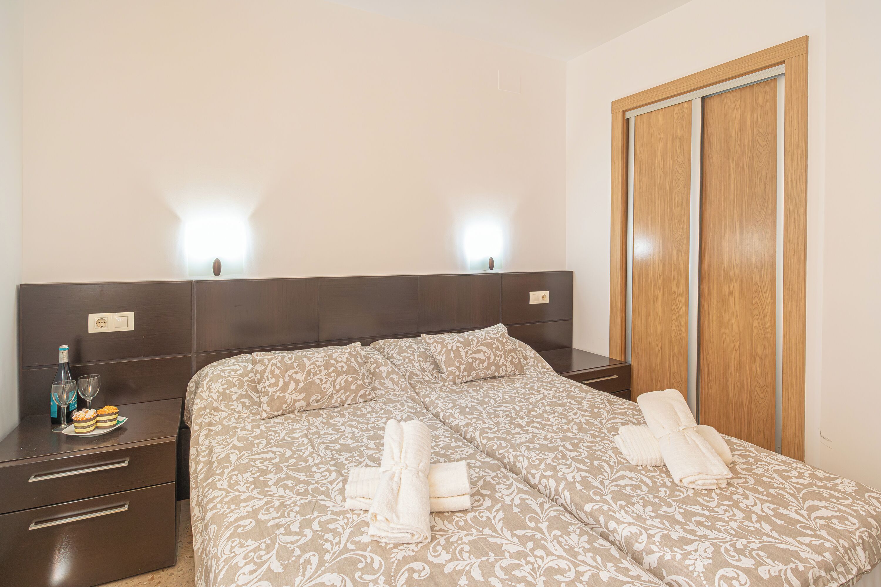 A neatly made bed with a patterned bedspread and two towels folded on it. There is a nightstand with a bottle and glasses on it, and a wooden wardrobe with a sliding door.