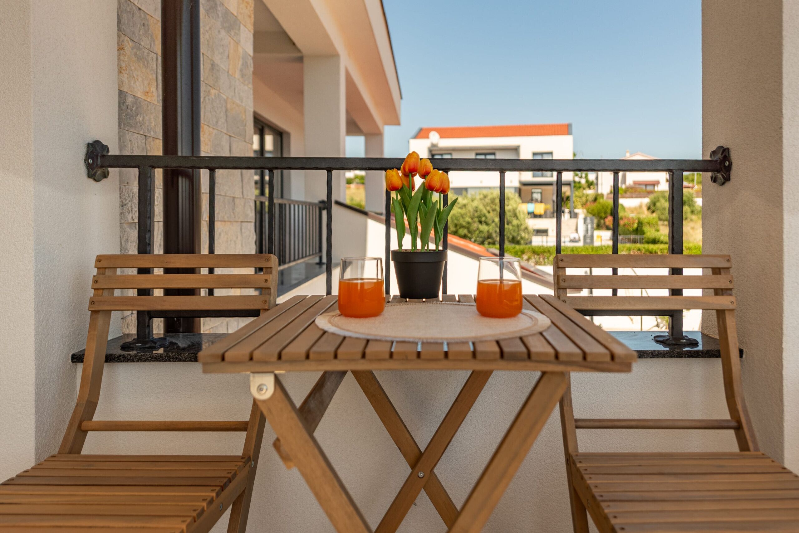 A wooden table with two chairs on a balcony, a potted plant with orange flowers, and a clear view of buildings in the background.