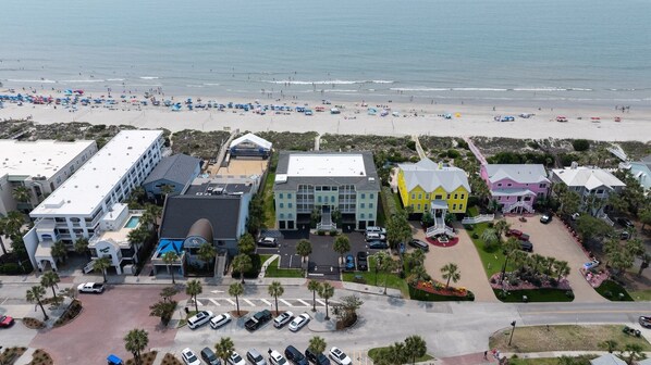 An aerial view of a coastal hotel with multiple buildings, a parking lot, and a beach in the background.