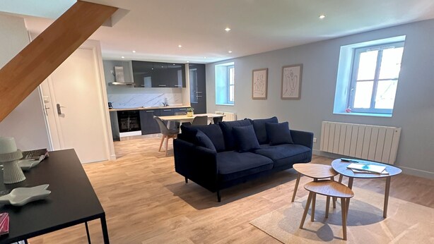 Una sala moderna con un sofá azul oscuro, una mesita redonda y una mesa de comedor. Se observa una cocina al fondo, con armarios oscuros y una ventana.