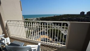 A balcony with white railings, two white lounge chairs, and a view of the beach and cityscape.