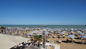 A beach with numerous umbrellas and sun loungers, a pier extending into the sea, and clear skies.