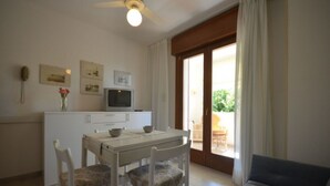 Une petite pièce bien éclairée, comprenant une table à manger, des chaises, un canapé, un ventilateur de plafond, un téléviseur et une porte-fenêtre coulissante donnant sur un espace extérieur.