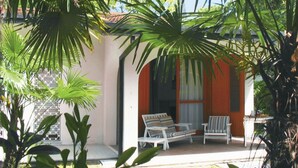 Un espace extérieur aménagé avec une zone de sièges couverte, comprenant deux chaises blanches et une table, le tout entouré d’une végétation tropicale luxuriante.