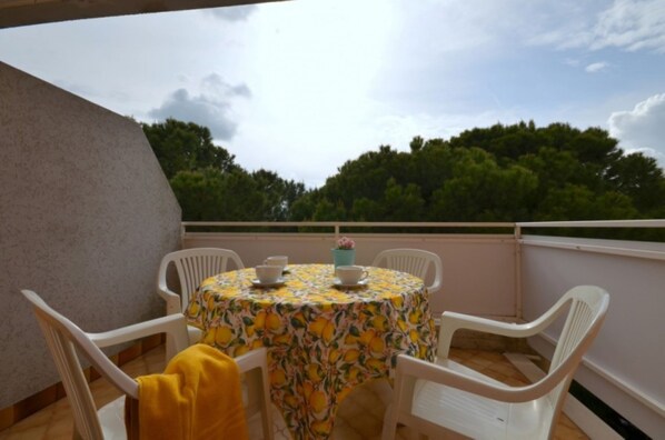 A round table with a floral tablecloth, surrounded by white chairs, on a balcony with a view of trees and a cloudy sky.