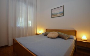A bedroom with a bed, two bedside lamps, a nightstand, a window with sheer curtains, and a wall-mounted picture.