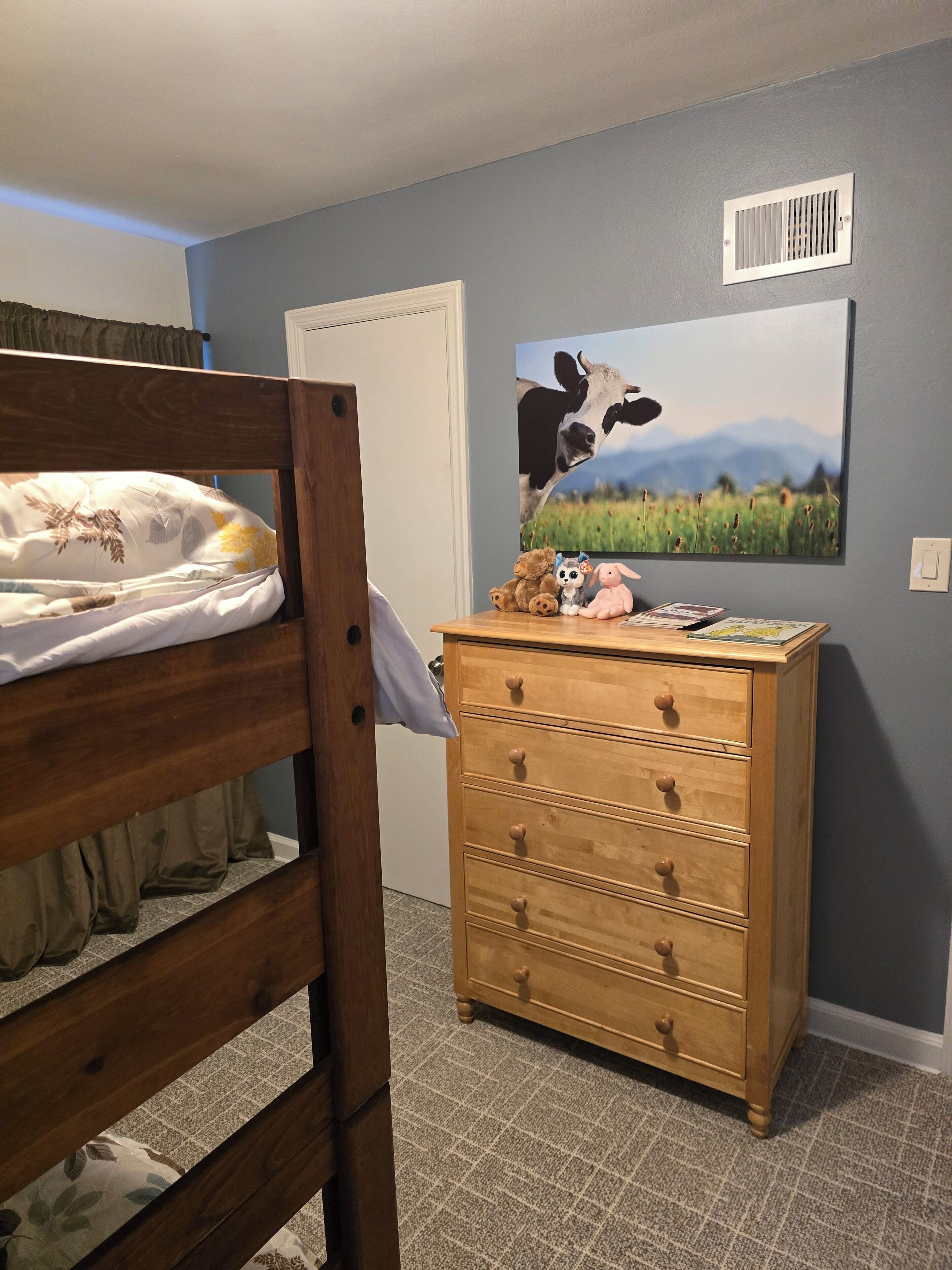 A bedroom with a bunk bed, a wooden dresser, a wall-mounted vent, and a landscape painting.