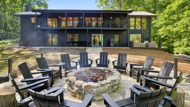 Una cabaña moderna de dos pisos con un hoyo de fuego de piedra rodeado de sillas Adirondack.
