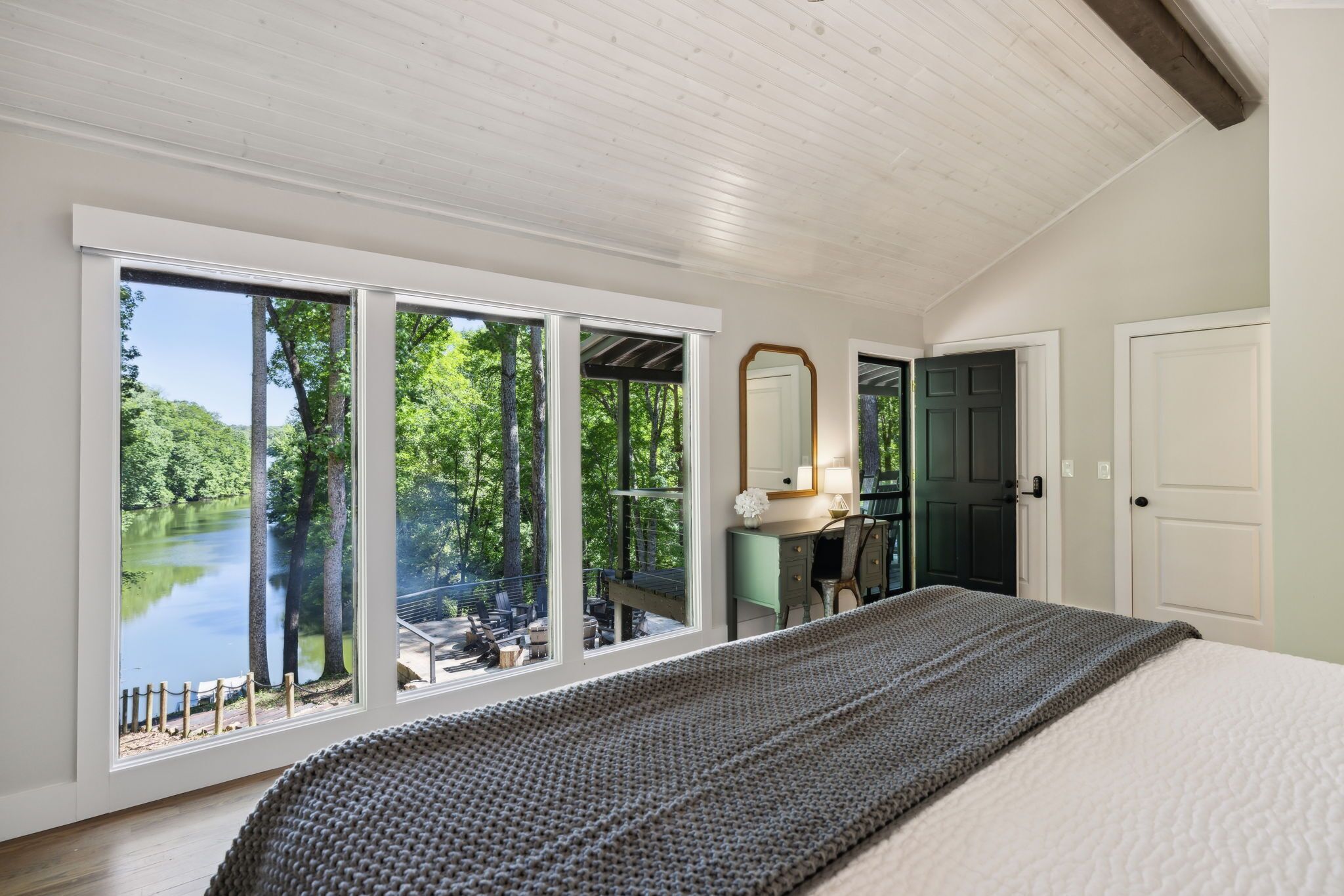 A bedroom with a large window overlooking a lake, a bed, a dresser, and a mirror.