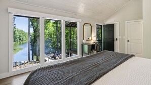 A bedroom with a large window overlooking a lake, a bed, a dresser, and a mirror.