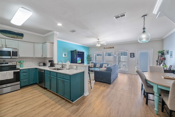 A modern kitchen with teal cabinets, a central island, and a dining area with a table and chairs. - Pet-Friendly Coastal Townhome | Heated Pool & Private Patio (Corpus Christi)