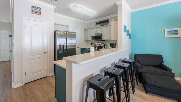 A modern kitchen with white cabinets, a stainless steel refrigerator, and a black countertop. There are four black stools at the counter. - Pet-Friendly Coastal Townhome | Heated Pool & Private Patio (Corpus Christi)