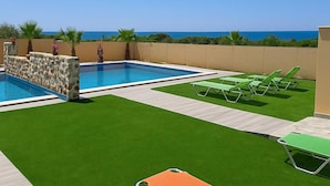 A swimming pool with a stone wall and palm trees, surrounded by green artificial grass and lounge chairs.