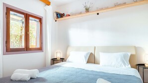 Een net opgemaakt bed met wit- en blauw beddengoed, een raam met gordijnen, en een plankje met decoratieve spullen.