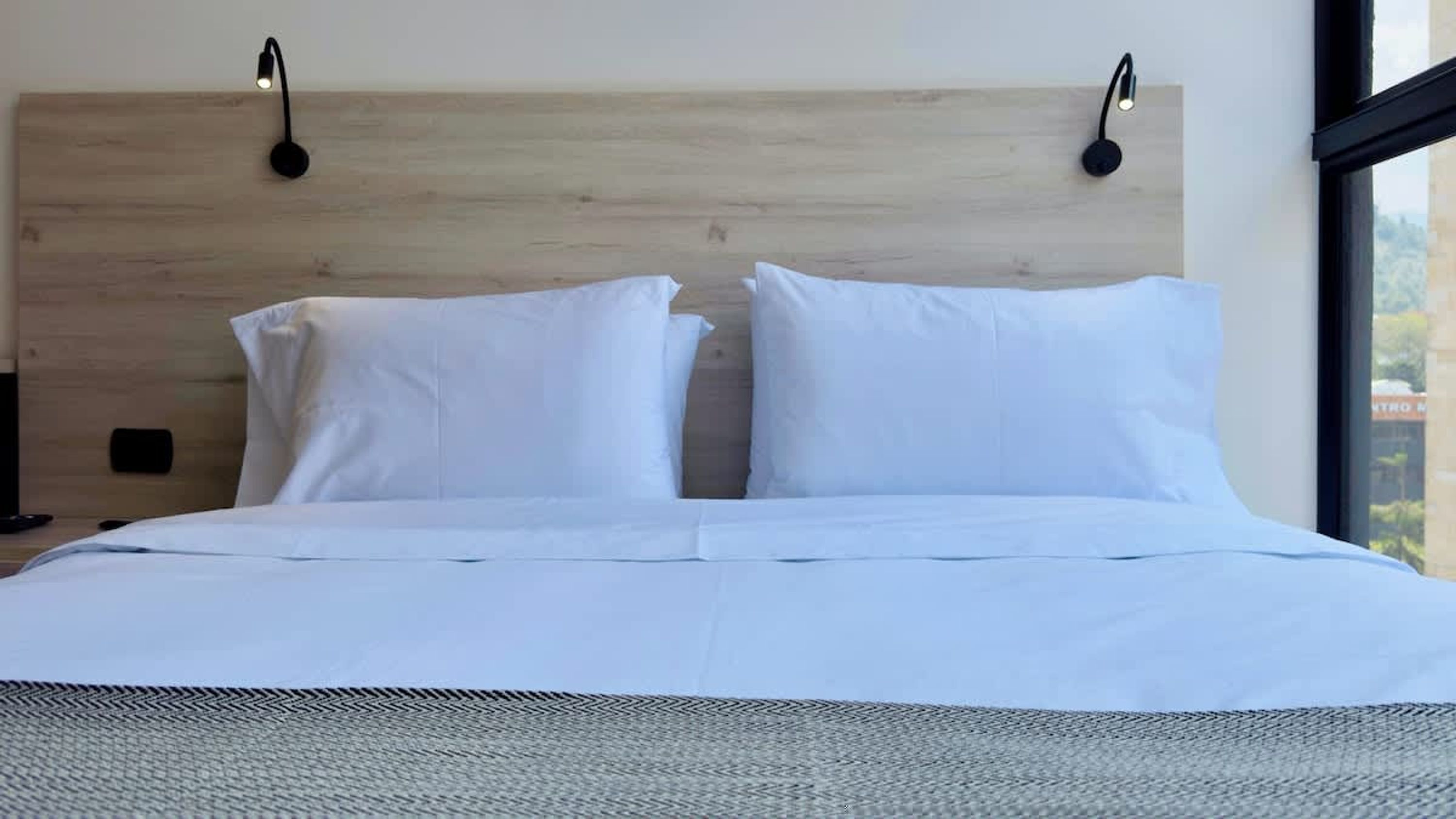 Una cama bien hecha con sábanas blancas, cabecera de madera y dos lámparas de lectura fijadas a la pared.