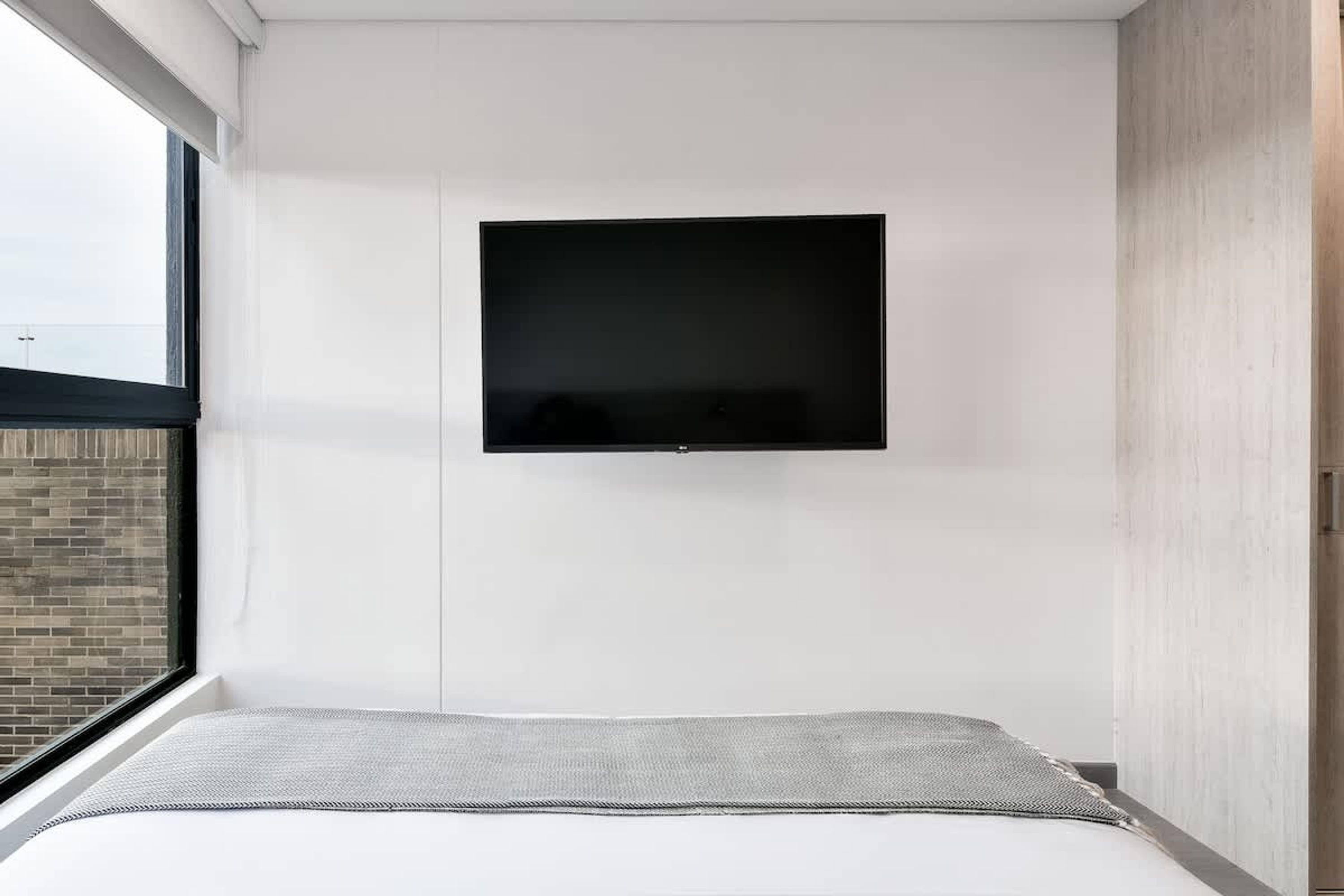 A modern bedroom with a bed, a mounted TV, and a window with a view of a brick building.