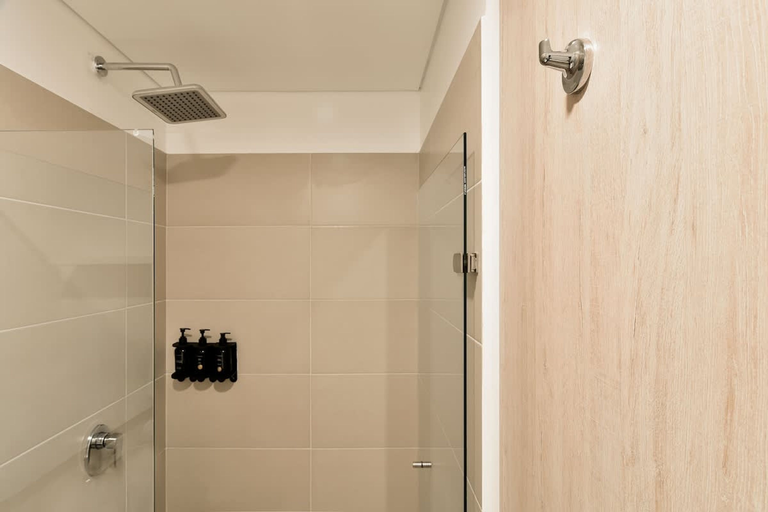 A modern bathroom with a glass shower enclosure, beige tiles, and a wooden door.