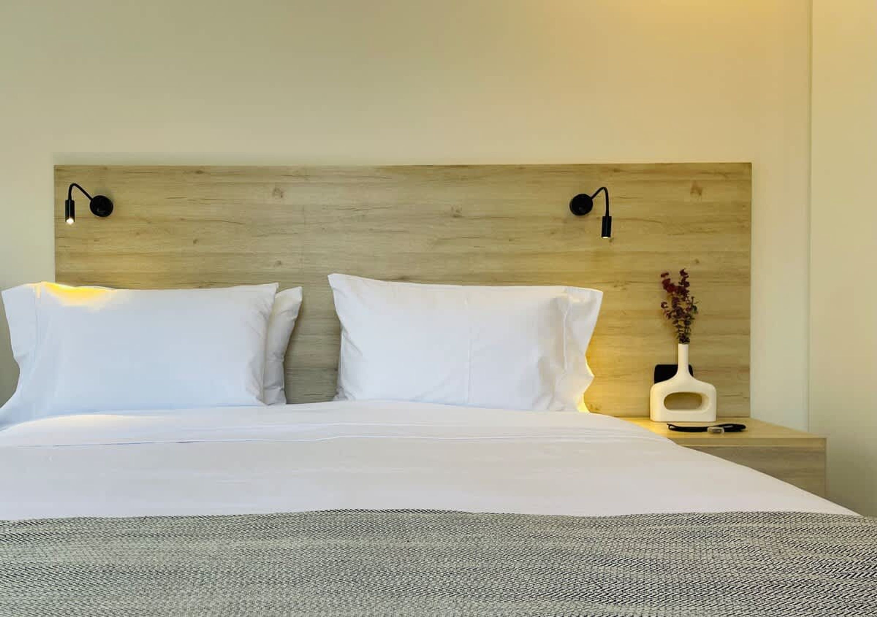 A neatly made bed with white linens and a gray throw blanket, a wooden headboard with two wall-mounted reading lights, and a bedside table with a white vase and a small plant.