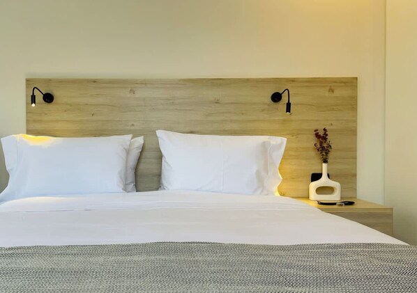 A neatly made bed with white linens and a gray throw blanket, a wooden headboard with two wall-mounted reading lights, and a bedside table with a white vase and a small plant.