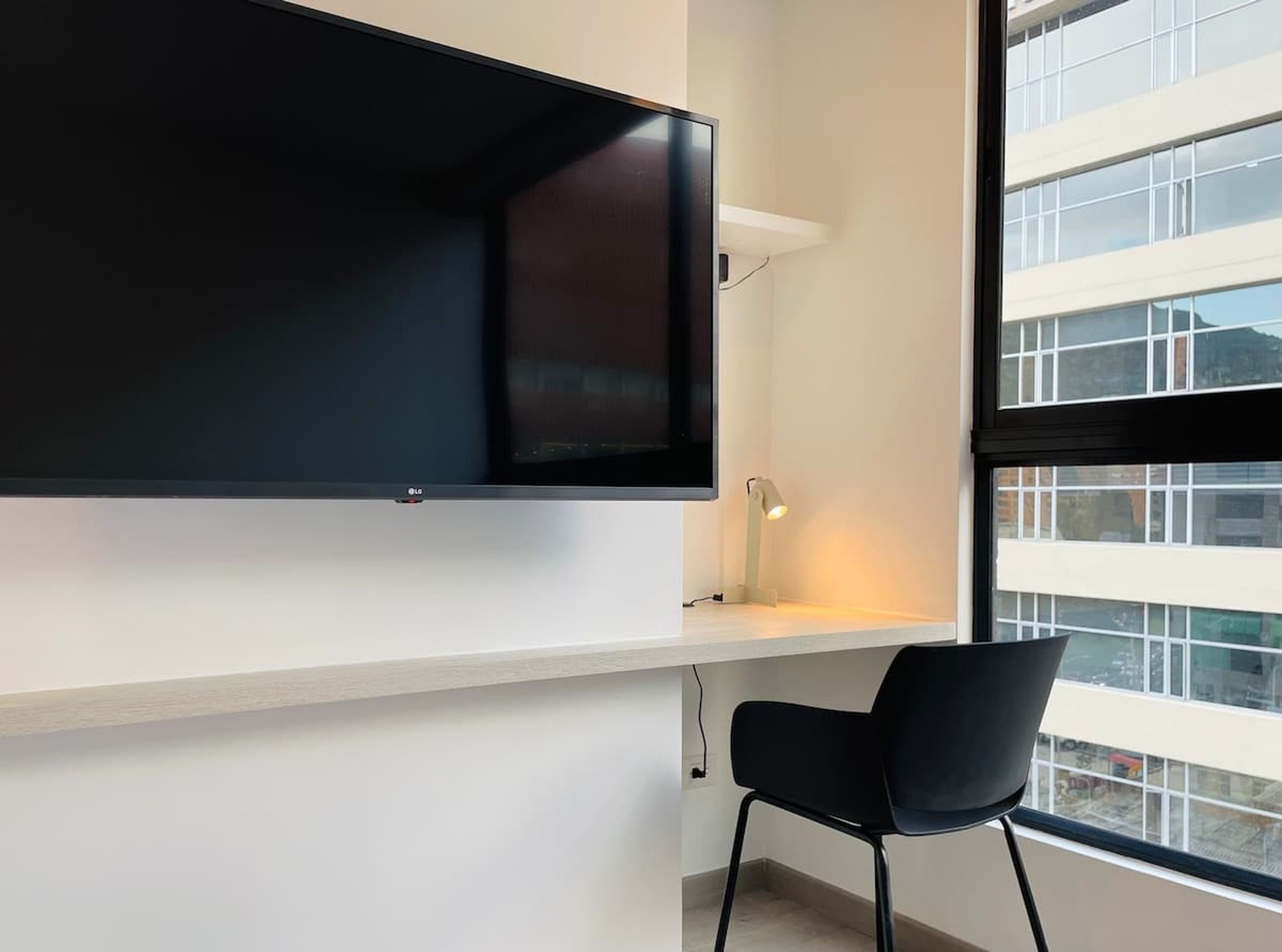 A modern room with a large flat-screen TV mounted on a white wall, a small desk with a lamp, and a black chair.