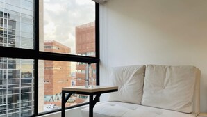 A room with a white sofa, a small table, and a large window offering a view of city buildings. - NOK Stylish Loft in Usaqu n (Bogotá)