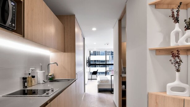 A modern kitchen with a built-in microwave, a sink, and wooden cabinets. There is a hallway leading to a dining area with a view of a building outside. - NOK Stylish Loft in Usaqu n (Bogotá)