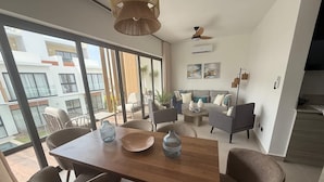 Un espace repas moderne avec une table et des chaises en bois, un ventilateur de plafond et de grandes fenêtres donnant sur l’extérieur.