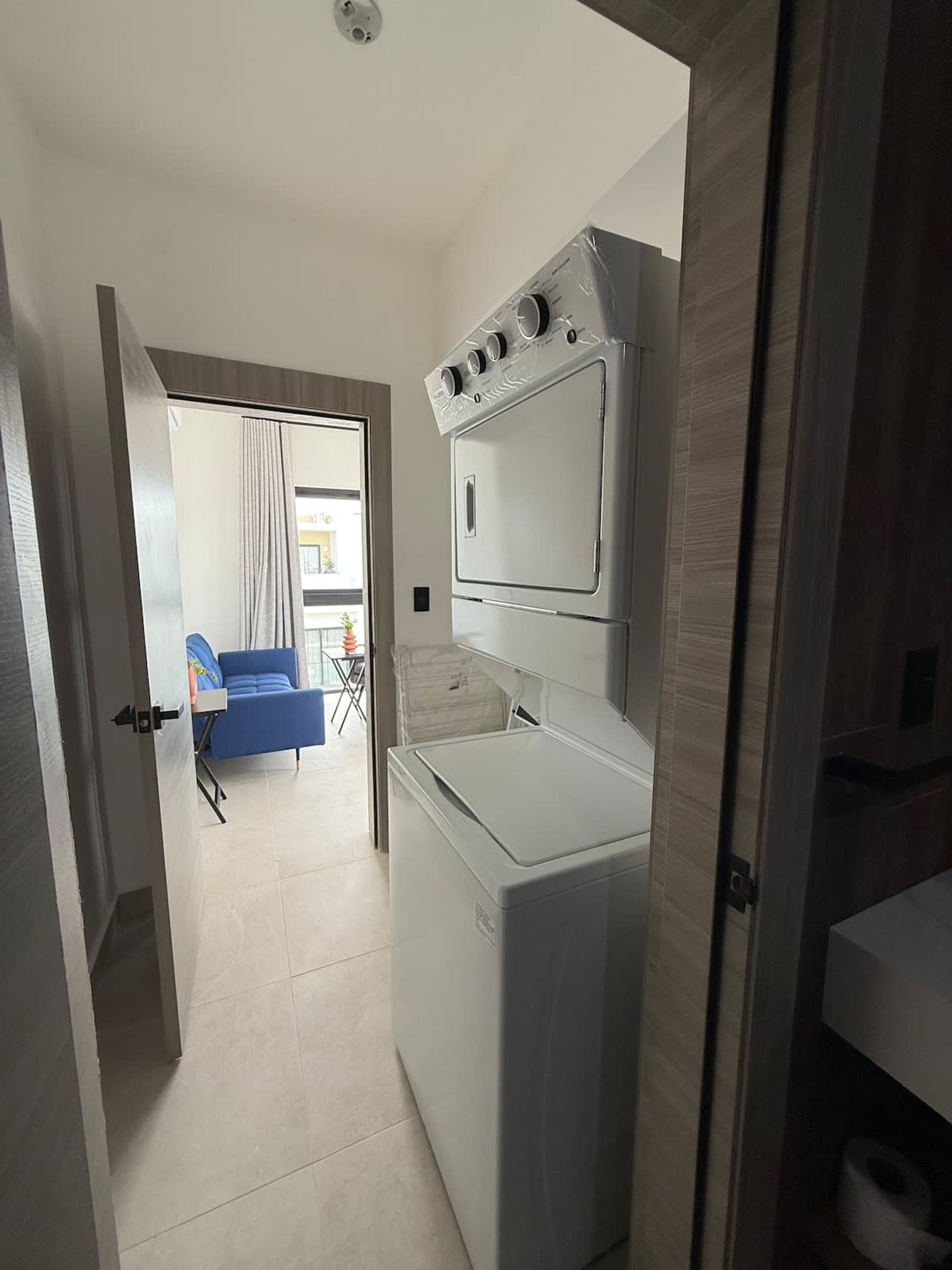 Una cucina compatta con lavatrice e asciugatrice integrate, un divano blu nella zona soggiorno adiacente e vista all'esterno tramite una porta vetrata.