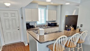 A kitchen with white cabinets, a granite countertop, a sink, a microwave, and a refrigerator. There are three white bar stools at the island. - Canal front on one side, bay on the other! Perfect midtown 2 bedroom condo next to Seacrets and close to everything! Wight Bay #452 (Ocean City)