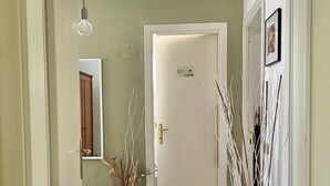 Un pasillo con una puerta blanca, un jarrón blanco con plantas secas, un tapete estampado y una lámpara.