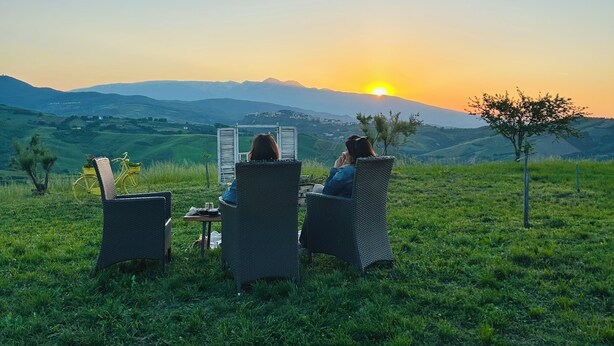 Tres sillas de mimbre mirando hacia una puesta de sol sobre un campo cubierto de hierba, con una cadena montañosa al fondo.