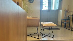 Una habitación acogedora con un sofá, una mesita y un banco. Hay una lámpara, una planta en maceta y un espejo en la pared. Una puerta da a otra habitación.