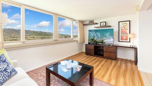 A living room with a large window, wooden floor, a glass coffee table, a sofa, a TV, and a desk with a plant. - Diamond Head & Golf Course Views at Island Colony w/Amazing Amenities (Honolulu)