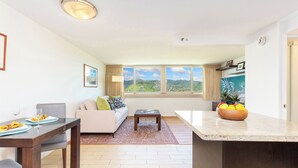 A modern living room with a dining area, large windows with a view of greenery, and a kitchen counter with a fruit bowl. - Diamond Head & Golf Course Views at Island Colony w/Amazing Amenities (Honolulu)